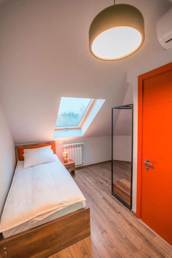 A bedroom with a skylight, a bed with white linens, a wooden floor, and a red door.