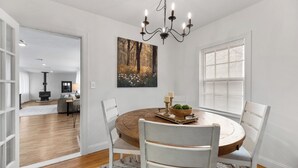 A dining area with a round wooden table, white chairs, a chandelier, and a painting of a forest scene. - Cozy vintage home in the heart of Riverside one block from golf, and much more (Wichita)