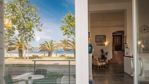 A view from a balcony overlooking palm trees and a beach, leading to a dining area with a table and chairs, and a kitchen with a refrigerator and a clock on the wall. - Hotel Il Gabbiano (Alghero)