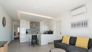 A modern kitchen with white cabinets, a central island, and a dining area with a grey sofa and yellow cushions. - Waterside Village - Sea View Apartment (Luz)