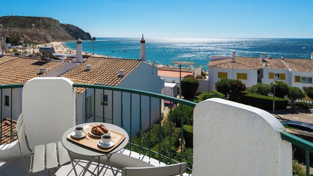 A balcony with a table set for two, overlooking a coastal town with buildings and a lighthouse. - Waterside Village - Sea View Apartment (Luz)