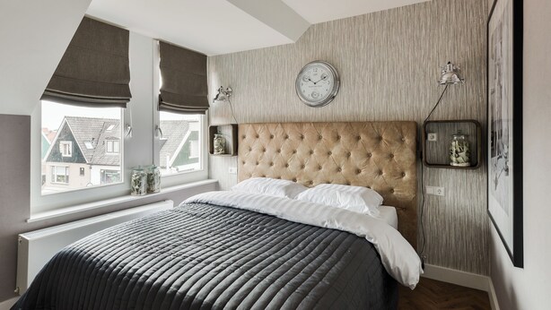 A bedroom with a tufted headboard, a large bed with a dark quilt, a wall clock, and a window with blinds. - Hotel aan de Haven (room 6) (Volendam)