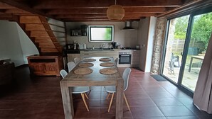 A dining area with a wooden table and chairs, a kitchen with a large window, and a staircase leading to an upper level. - L'escale - Longère en Campagne 6 Voyageurs, 3 Chambres (Pouldreuzic)
