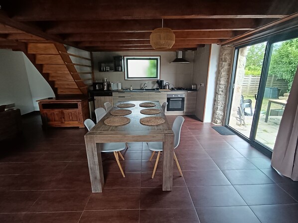Un comedor con una mesa y sillas de madera, una cocina con un ventanal grande y una escalera que lleva a un nivel superior.