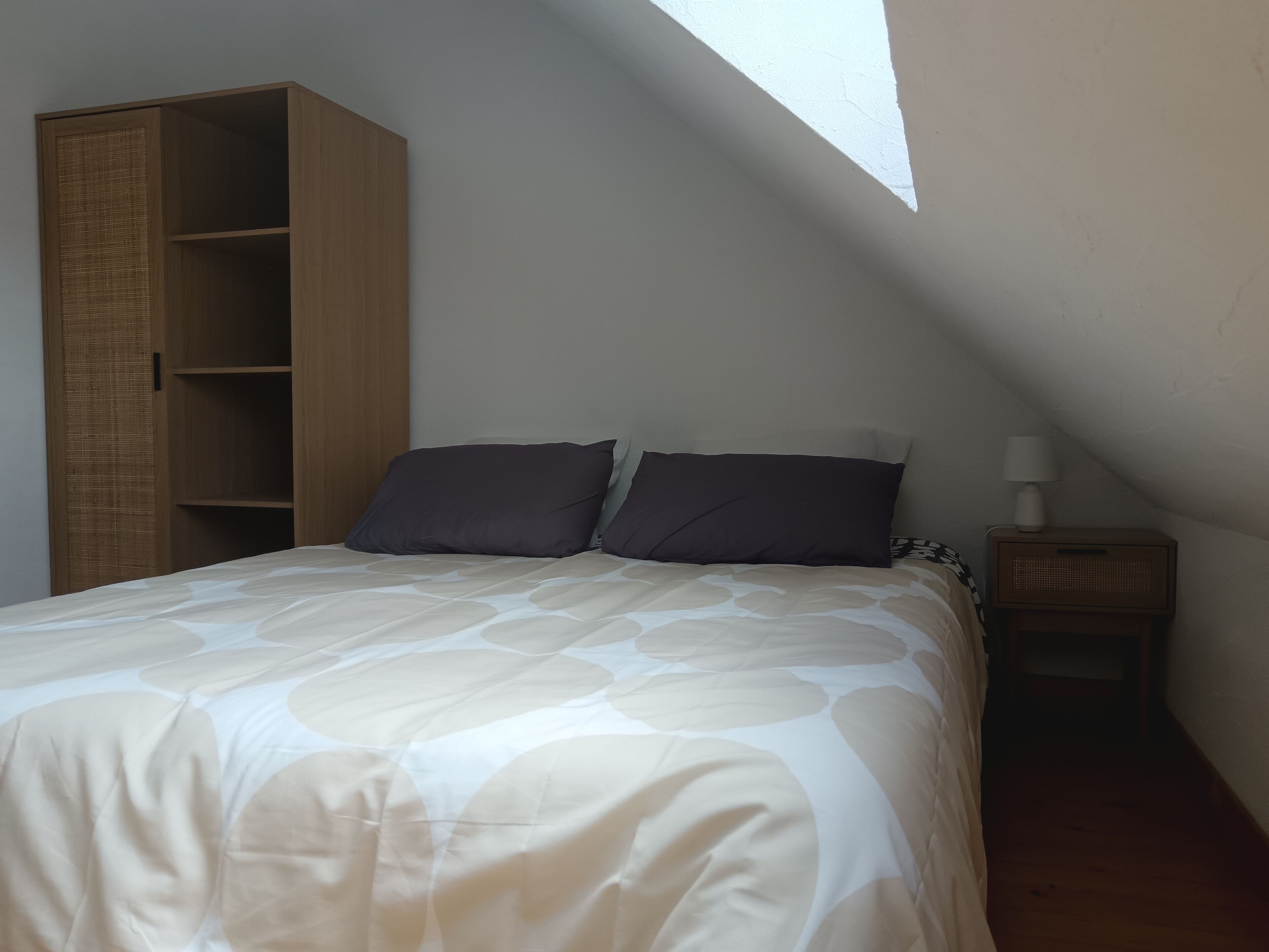 Une chambre à coucher avec un lit, deux oreillers, une table de chevet avec une lampe, une armoire en bois et un plafond en pente avec une lucarne.