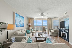 A spacious living room with a large sofa set, a coffee table, and a fireplace. There is a balcony with a view of the ocean and a ceiling fan.