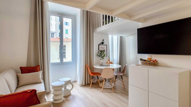 A modern living room with a sofa, a dining table with chairs, a flat-screen TV, and a large window with curtains. - SUEDE PROMENADE – Duplex apartment at 1 min from the sea, 2 bathrooms (Nice)