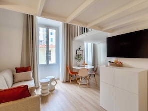 A modern living room with a sofa, a dining table with chairs, a flat-screen TV, and a large window with curtains.