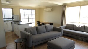 Una sala moderna con un sofá gris, un reposapiés y una pequeña mesa. Se observa una zona de cocina con una mesa de comedor y sillas, además de un ventanal con cortinas.