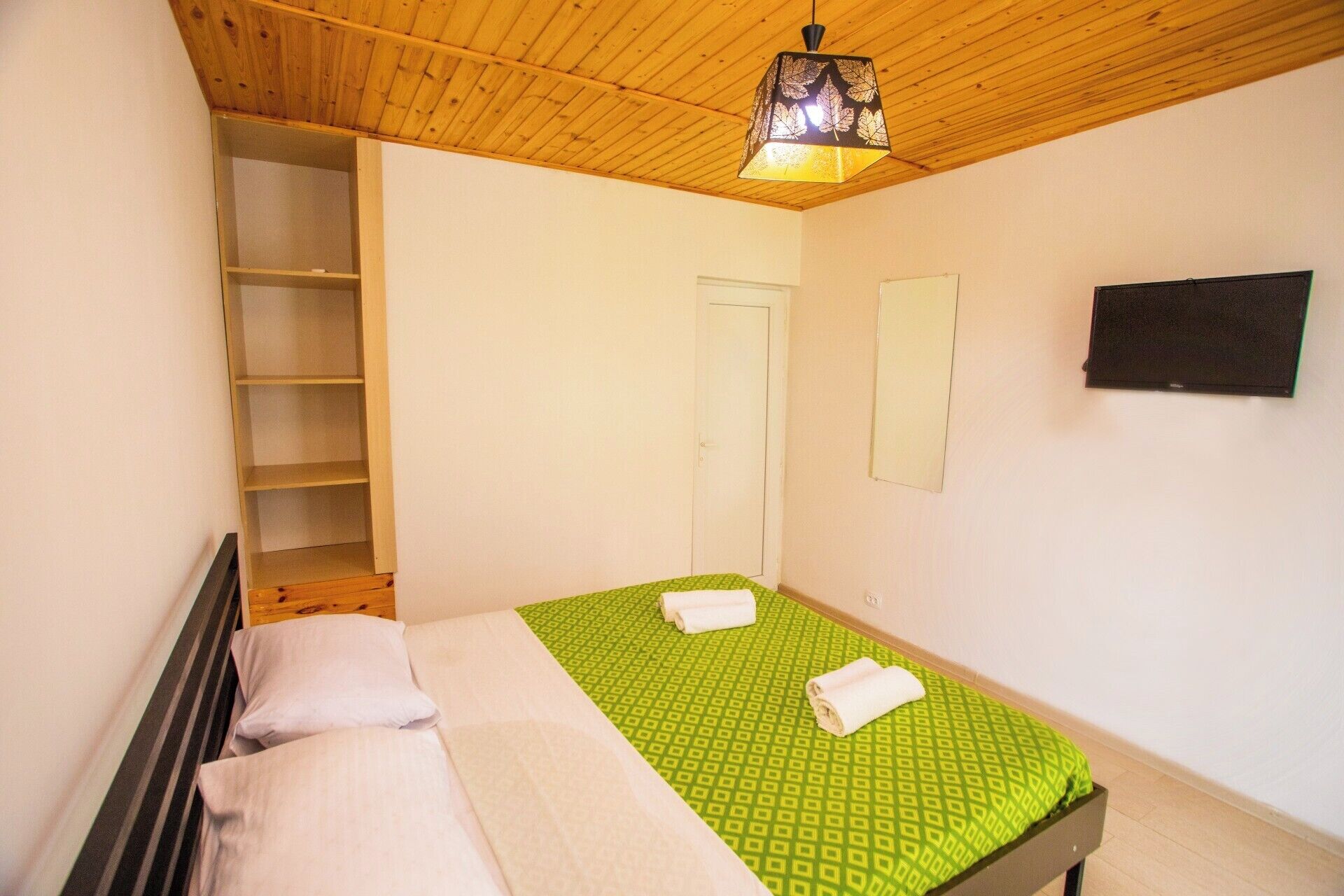 A bedroom with a wooden ceiling, a bed with green bedding, a white door, and a small black object on the wall.