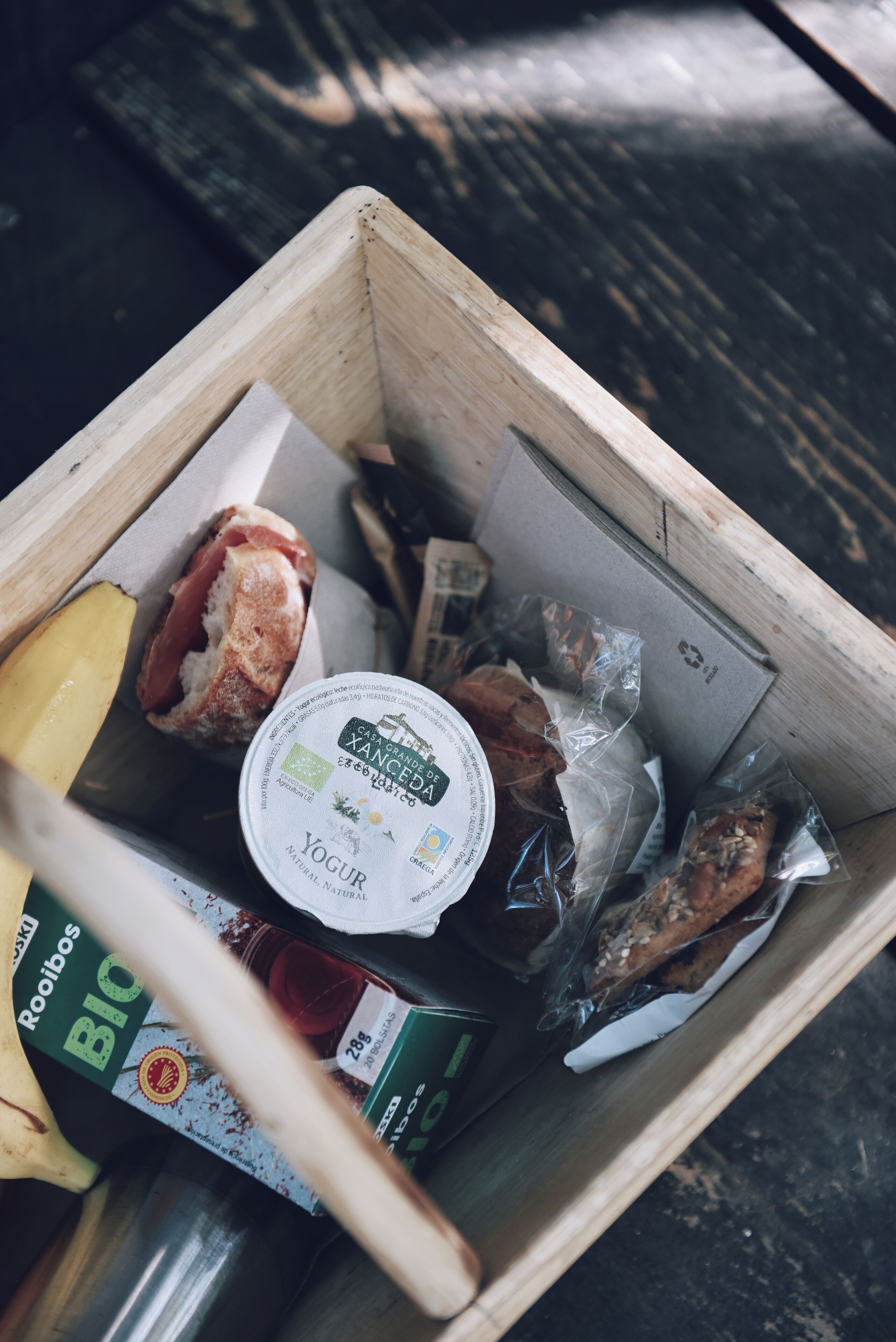 Una caja de madera llena de diversos alimentos, como una barra de pan, un recipiente con yogur y aperitivos envasados.
