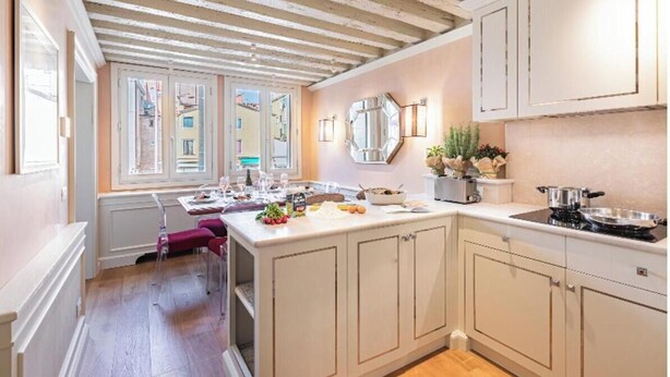 Una cocina con una isla central, armarios blancos, techo de madera y vista a la calle a través de las ventanas.