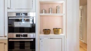 Una cocina con armarios blancos, horno empotrado y una estantería con objetos decorativos.