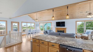 Cocina amplia con armarios de madera, encimera de granito y lavavajillas integrado. Cuenta con comedor con sillas blancas y una mesa, además de sala de estar con sofá y chimenea. Amplias ventanas ofrecen vistas del entorno.