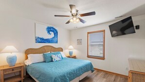 Une chambre à coucher comprenant un lit, deux tables de chevet, un ventilateur de plafond, une télévision et une fenêtre avec des stores.