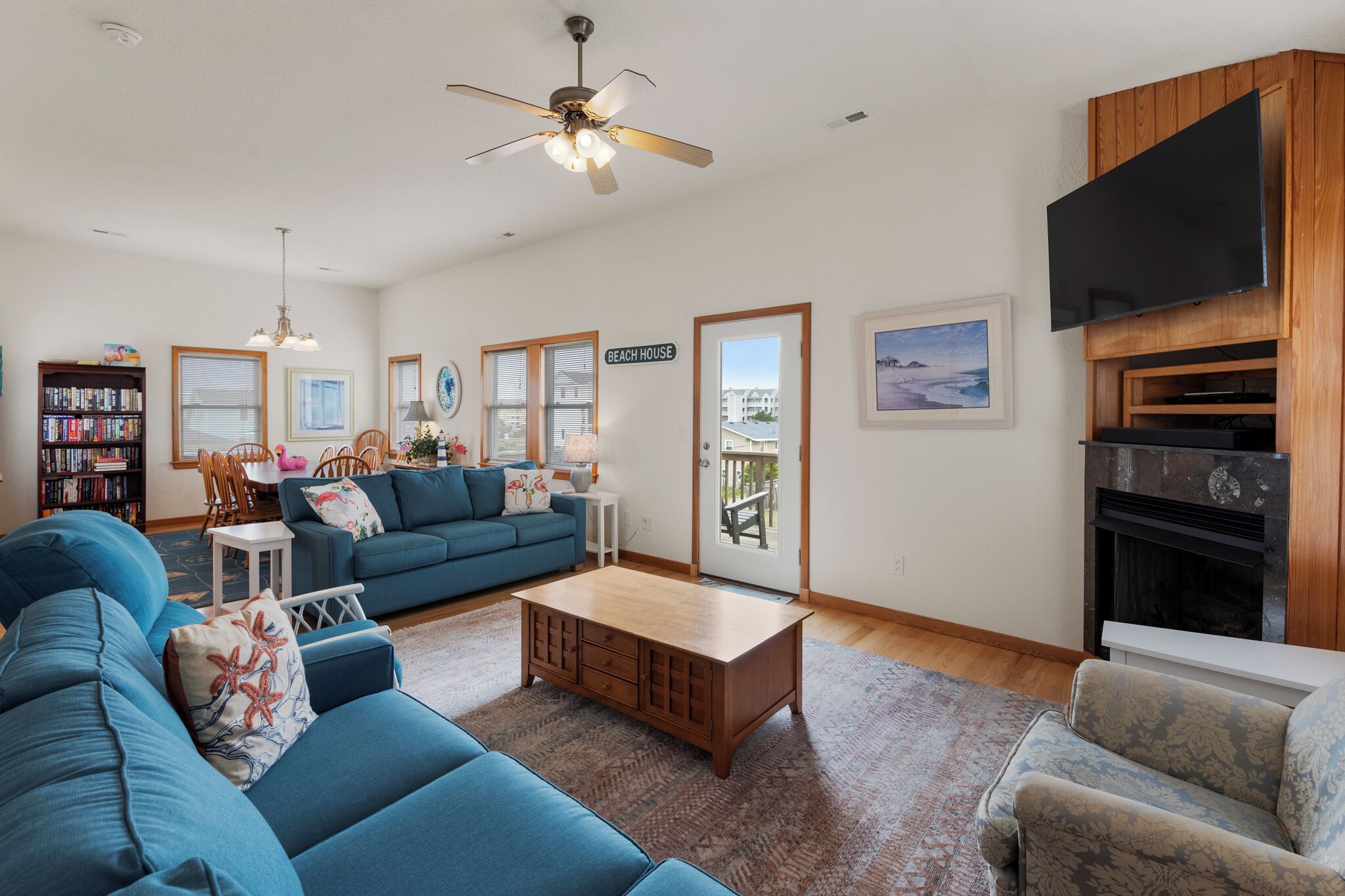 A living room with a blue sofa, wooden coffee table, and a fireplace. There is a bookshelf, a dining table, and a ceiling fan.