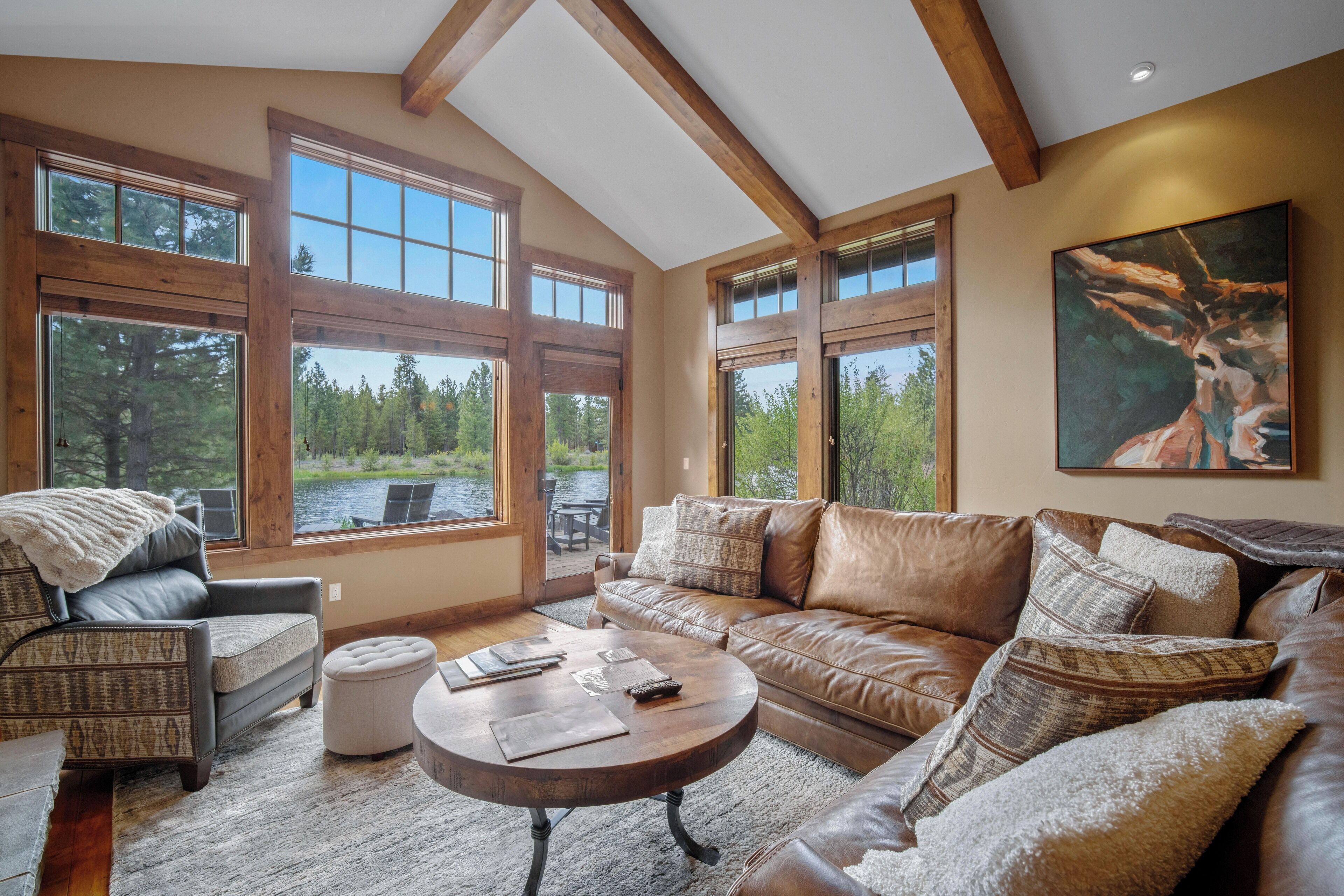 A spacious living room with a large window offering a view of a lake and forest, a leather sofa, a wooden coffee table, and a patterned armchair.
