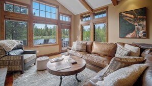 A spacious living room with a large window offering a view of a lake and forest, a leather sofa, a wooden coffee table, and a patterned armchair. - Lake View Home with Access to Exclusive Golf (Bend)