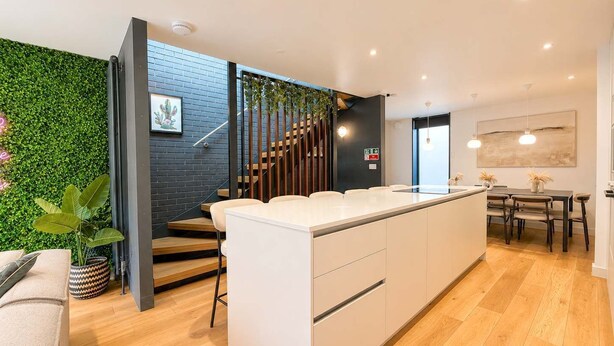 A modern kitchen with a central island, wooden flooring, and a staircase with a green wall.
