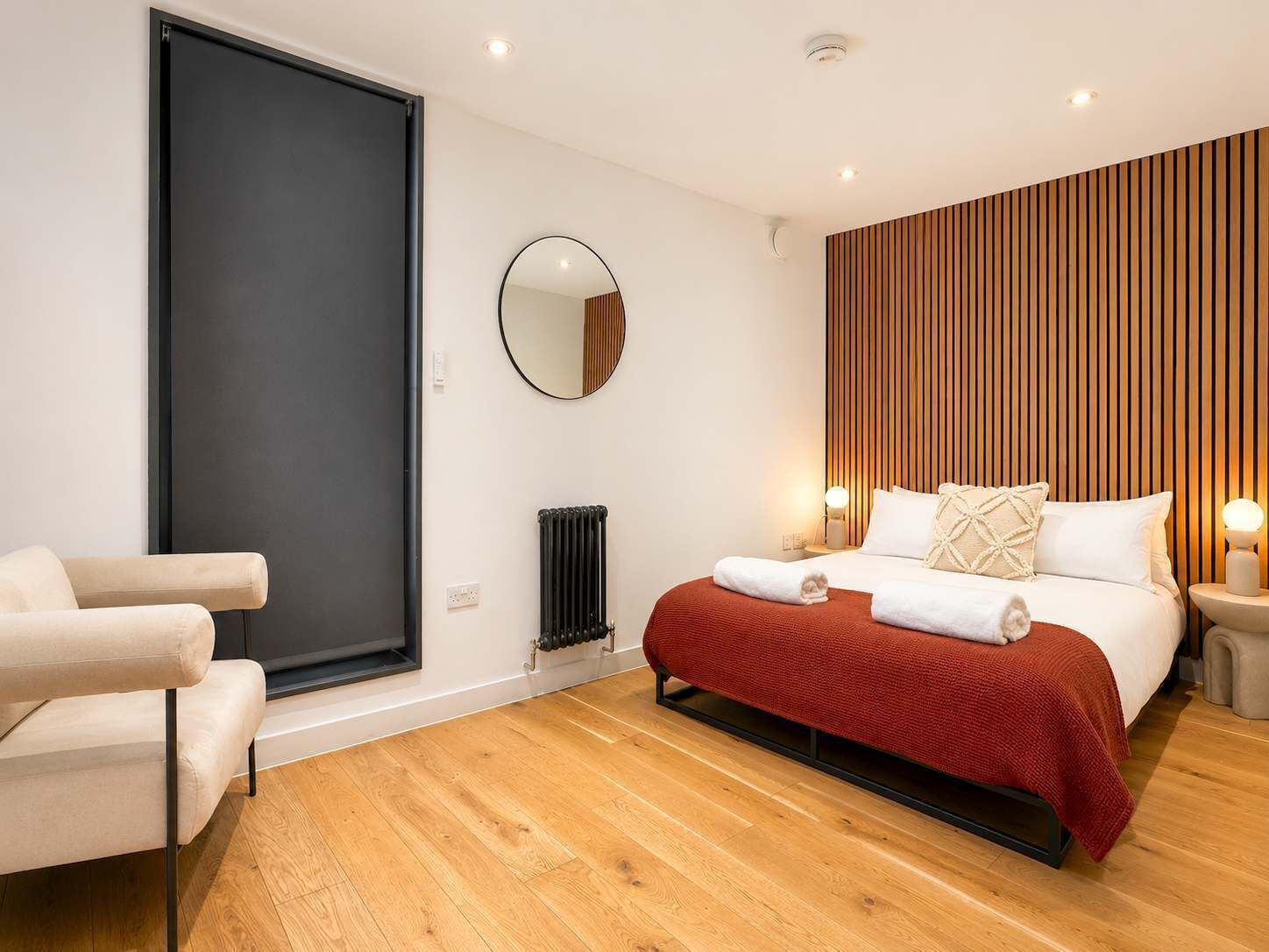 Ein modernes Schlafzimmer mit einem Bett, einem Sessel, einem Heizkörper, einem Spiegel und einer Wandpaneele aus Holz.