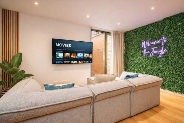 Una sala de estar moderna con un televisor de pantalla plana grande mostrando una selección de películas, un letrero de neón que dice 'Welcome to the Jungle' y una pared verde.