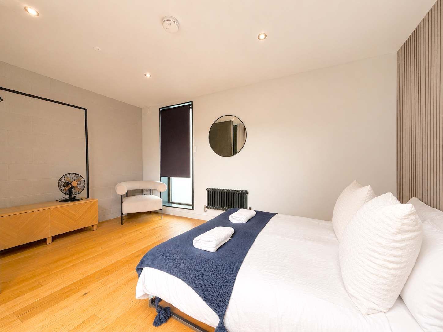 A modern bedroom with a bed, a chair, a radiator, and a wall-mounted mirror.