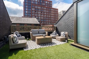 Eine Terrasse mit Gartenmöbeln, Kunstrasen und Blick auf ein hohes Gebäude.