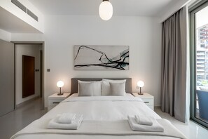 Ein modernes Schlafzimmer mit einem großen Bett, zwei Nachttischen mit Lampen, einem an der Wand hängenden abstrakten Gemälde und Blick auf ein Gebäude durch ein Fenster.