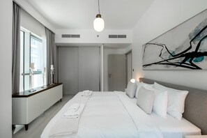 A modern hotel room with a large bed, a bedside table, a window with curtains, and a wall-mounted abstract painting.