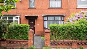 A red brick house with a front door, two windows, and a small garden with purple flowers. - Host & Stay | Broadway (Chadderton)