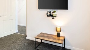 A hallway with a white door, a black wall-mounted TV, a small shelf with a plant, and a table with a lamp. - Host & Stay | Broadway (Chadderton)