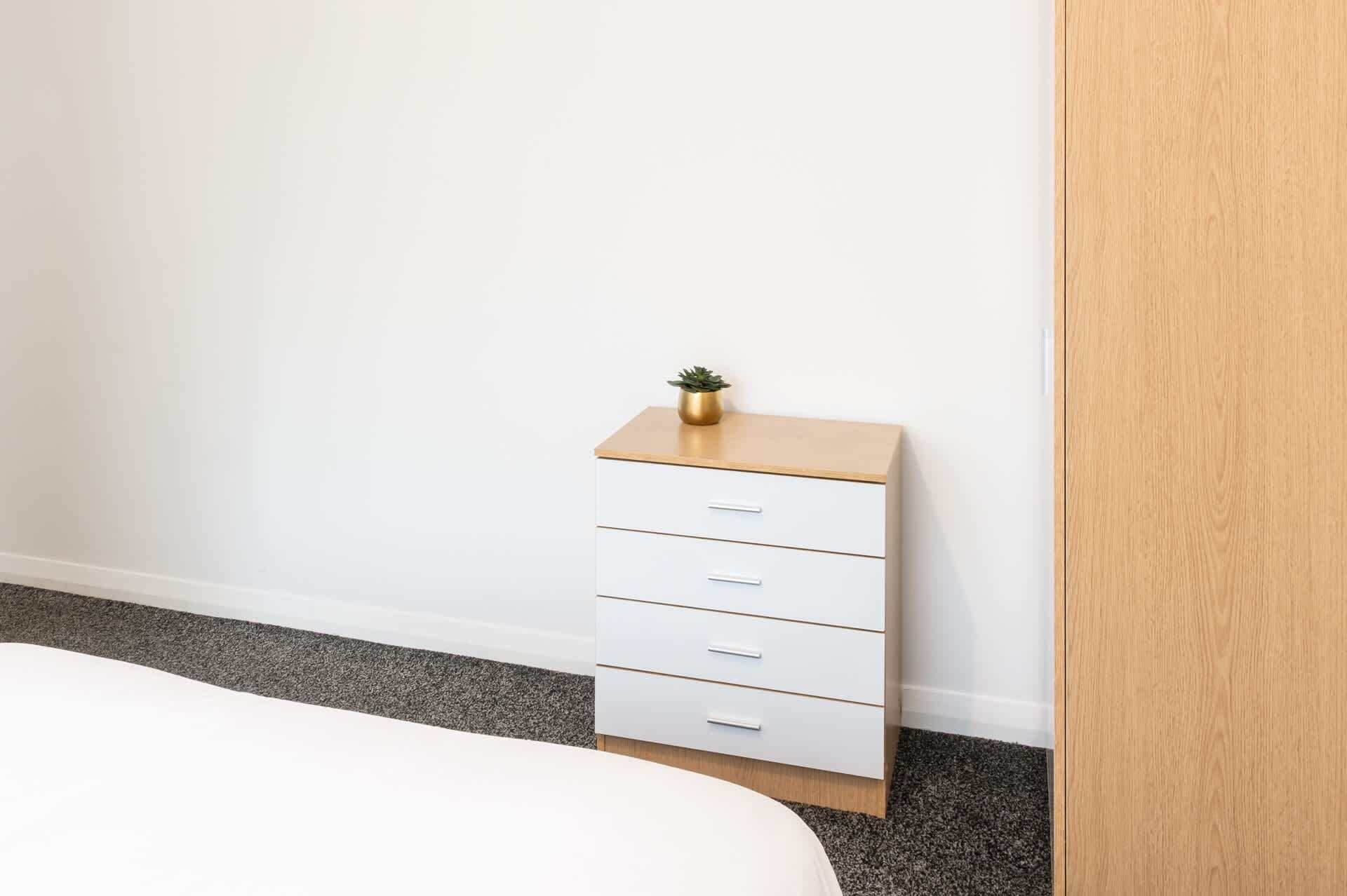 Un dormitorio con una cama, un aparador blanco con cajones, una planta pequeña encima y una puerta de madera.