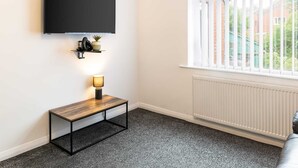A room with a mounted TV, a small table with a lamp, a radiator, and a window with blinds. - Host & Stay | Broadway (Chadderton)