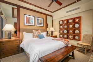 A hotel room with a large bed, wooden headboard, two framed pictures, a wall of decorative shells, a wooden bench, a wicker chair, and a ceiling fan.