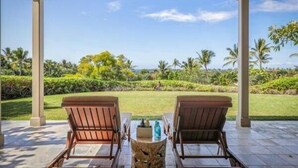 Un espace extérieur abrité, aménagé avec deux chaises en bois et une petite table, offrant une vue sur un jardin bien entretenu, agrémenté de palmiers et surplombant un ciel dégagé.