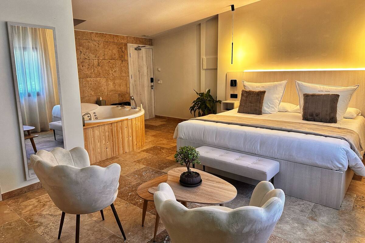 A modern hotel room with a large bed, a sitting area with two chairs, a small table, a bathtub, and a potted plant.