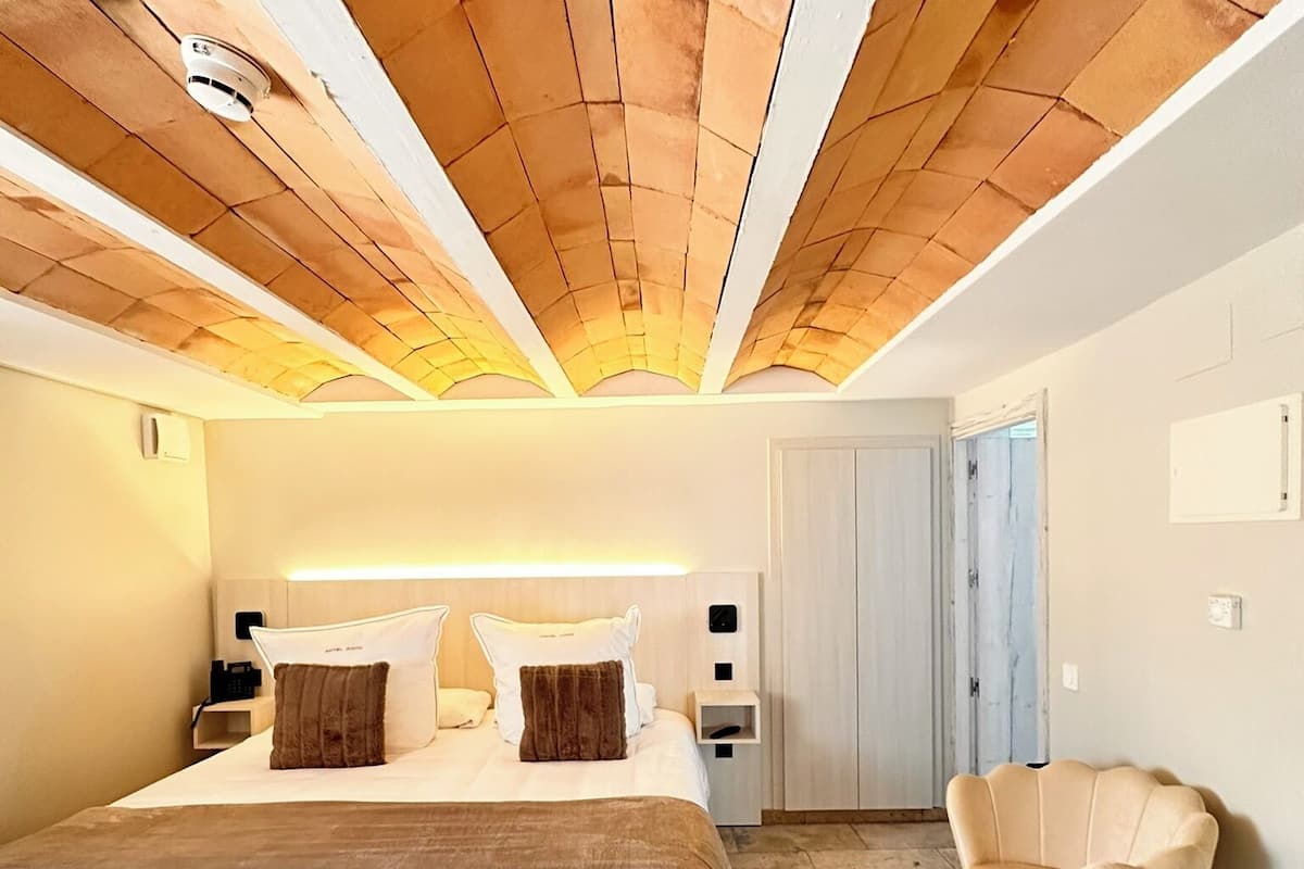 A bedroom with a large bed, a wooden ceiling, a nightstand, a chair, and a door.