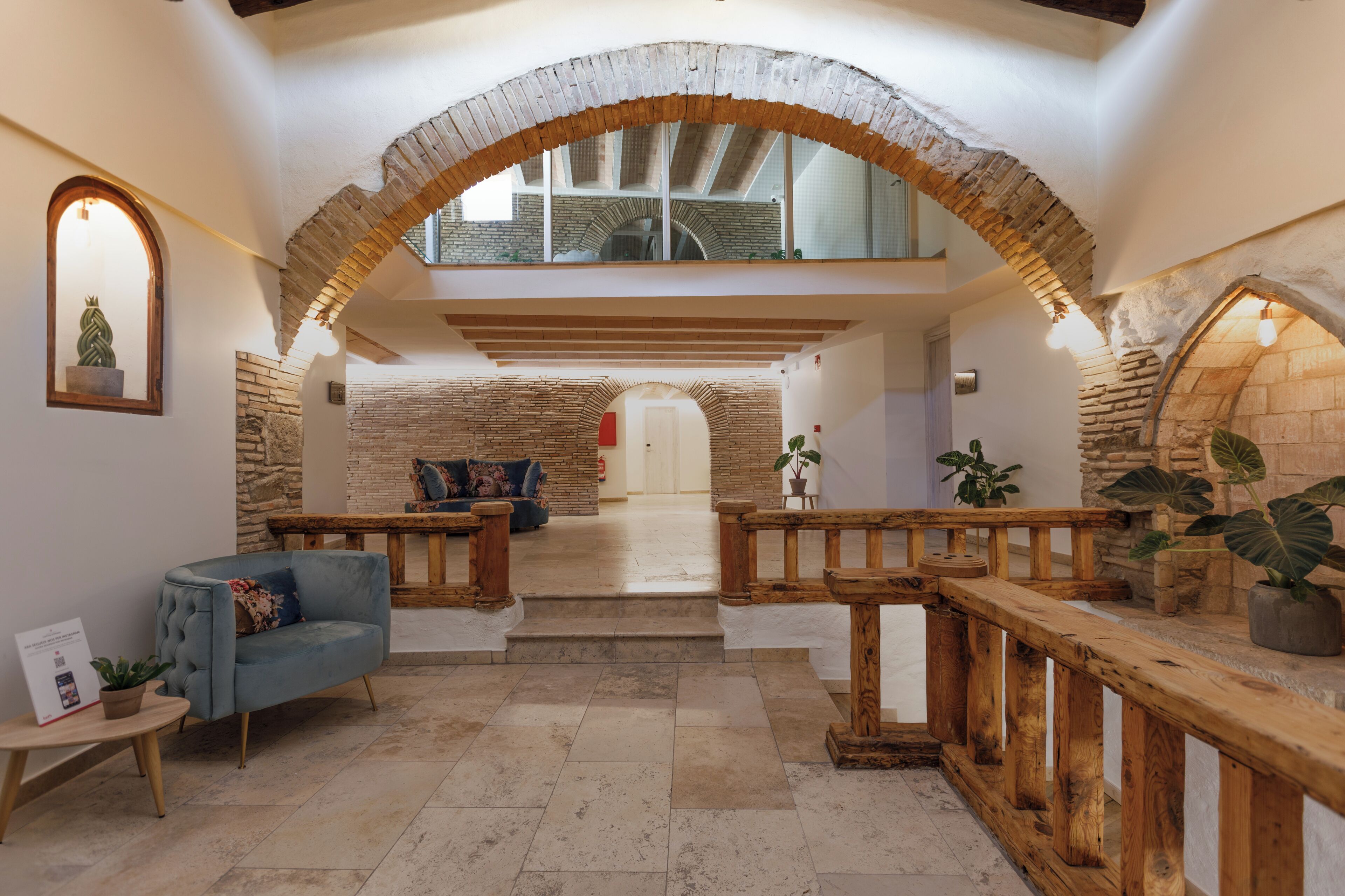 An interior with a stone archway, a sofa, wooden railings, and a staircase leading to a second floor.