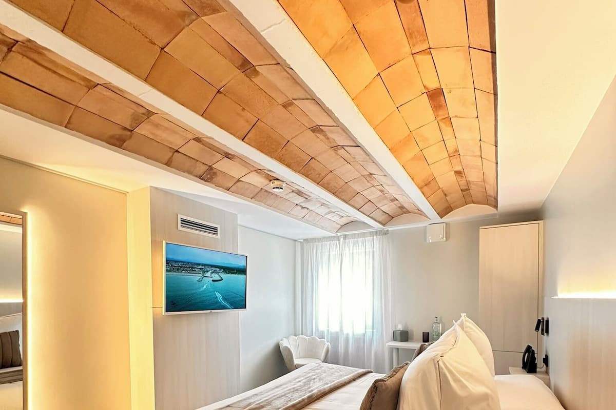A modern hotel room with a large bed, a flat-screen TV, a wooden ceiling, and a view of the sea.