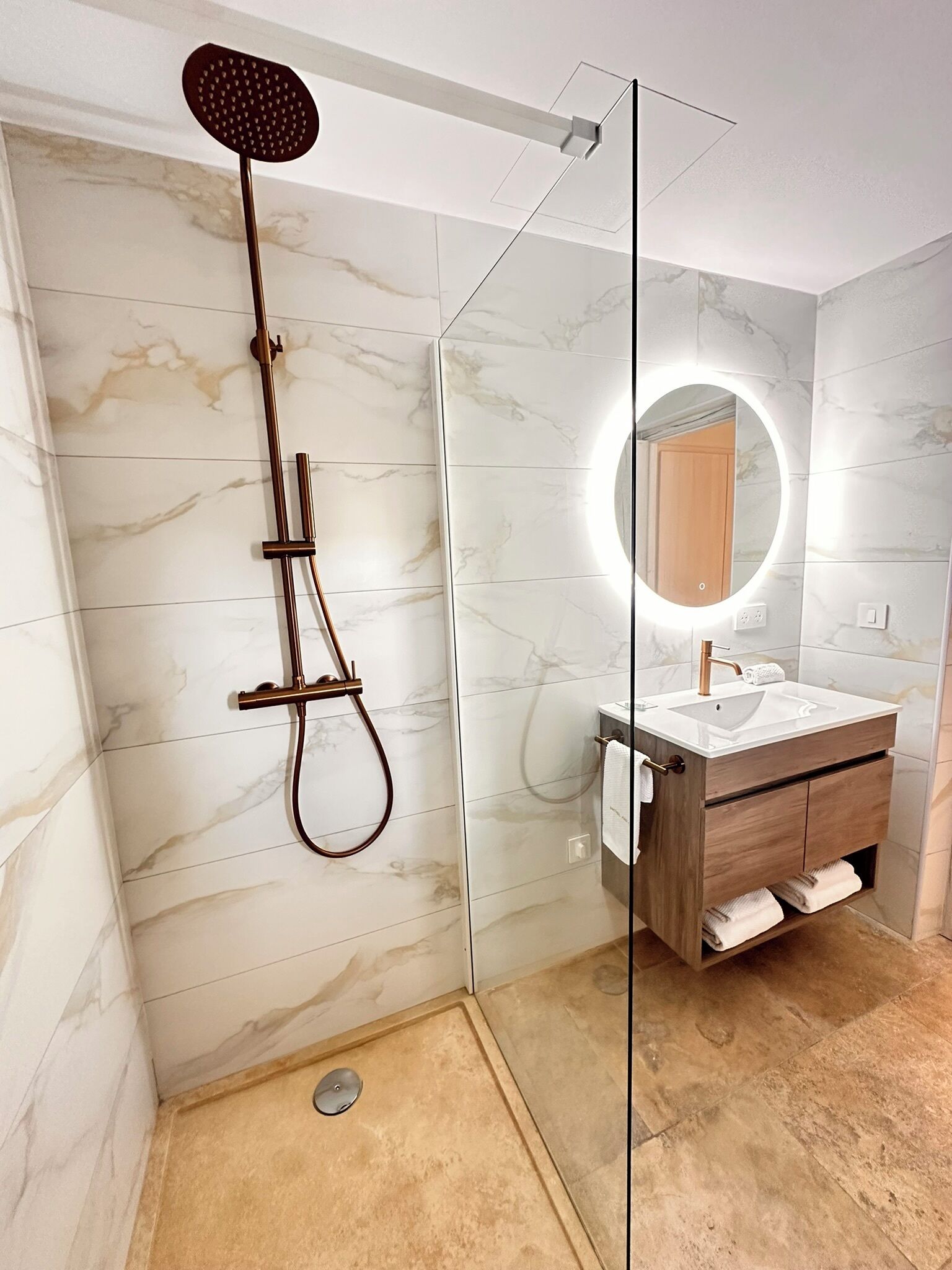 A modern bathroom with a glass-enclosed shower, a vanity with a marble countertop, and a round mirror with integrated lighting.