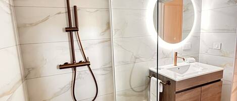 Un baño moderno con ducha envuelta en vidrio, tocador de mármol y espejo redondo con iluminación integrada.