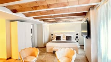 A modern hotel room with a large bed, two armchairs, a wooden table, and a TV on the wall.