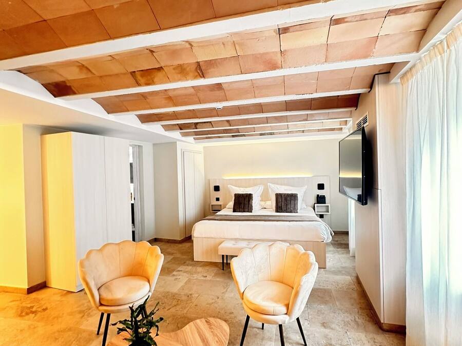 A modern hotel room with a large bed, two armchairs, a wooden table, and a TV on the wall.