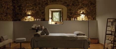 Una habitación con pared de piedra, una cama con almohadas, una mesita, una lámpara y un estante con toallas.