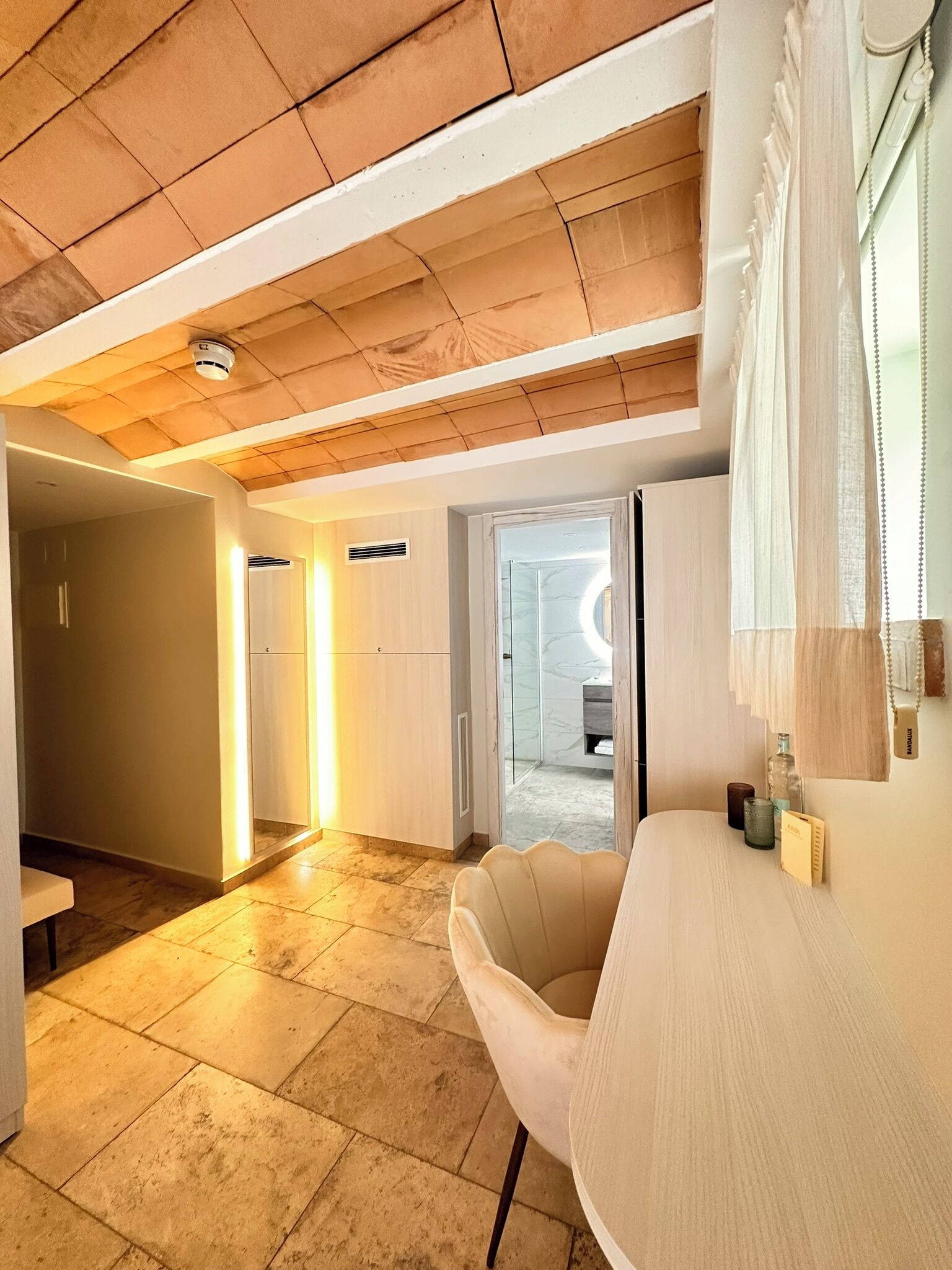 A modern interior with a wooden ceiling, a round table, and a chair. There is a hallway leading to a bathroom with a glass door.