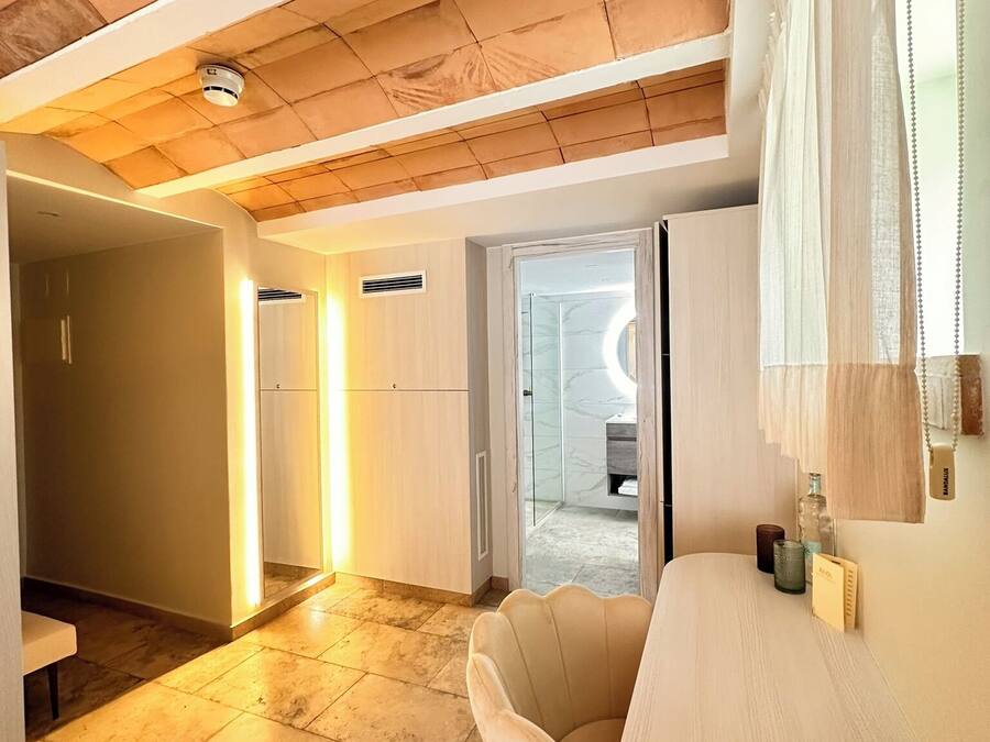 A modern interior with a wooden ceiling, a round table, and a chair. There is a hallway leading to a bathroom with a glass door.