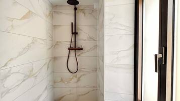 Un baño moderno con paredes de mármol, ducha con cabezal de ducha manual, una toalla en el suelo y artículos de aseo en un repisa.