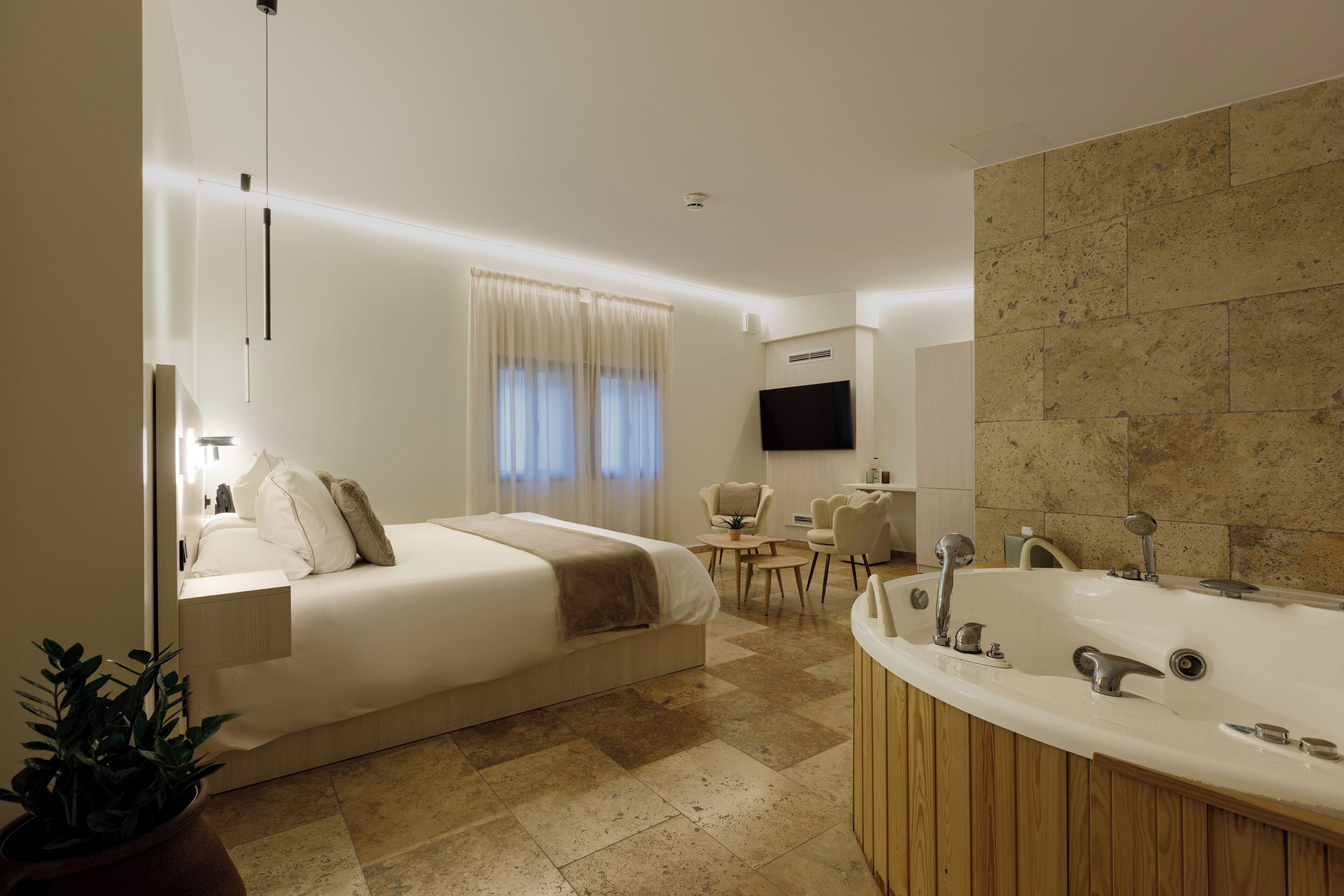 A modern hotel room with a large bed, a sitting area with a table and chairs, a large bathtub, and a flat-screen TV mounted on the wall.