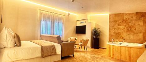A modern hotel room with a large bed, a seating area with chairs, a small table, a TV, and a bathroom with a bathtub and sink.