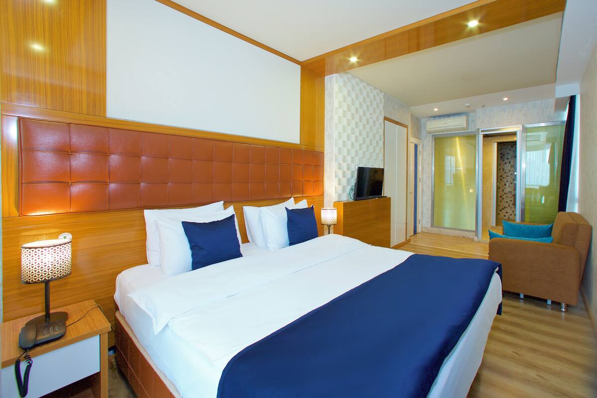 Una camera d'albergo con un letto grande, un comodino con una lampada, una televisione e un'area salotto con una poltrona.